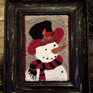 May include: A framed needlepoint artwork featuring a snowman with a red hat, black top hat, and a red and black striped scarf. The snowman is against a beige background.