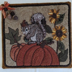 May include: A brown, tan, and green needlepoint rug featuring a squirrel perched on a pumpkin. The squirrel is grey and has a bushy tail. The pumpkin is orange and has a green stem. Sunflowers are depicted in the background.