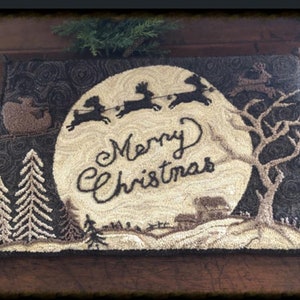 Puede incluir: Una alfombra de punto de aguja de color marrón y crema con una escena de Santa Claus y sus renos volando por el aire frente a una luna llena. La luna tiene las palabras "Merry Christmas" escritas en ella. La alfombra está rodeada por un borde marrón oscuro.