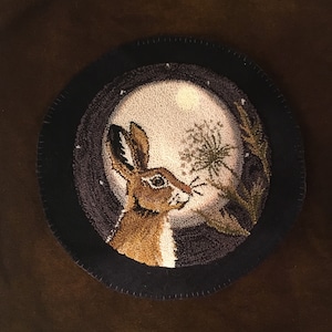 Puede incluir: Una alfombra redonda de fieltro negro con un borde cosido. El centro presenta una luna blanca y esponjosa con un conejo marrón mirando un diente de león. El conejo está en primer plano y el diente de león en el fondo.