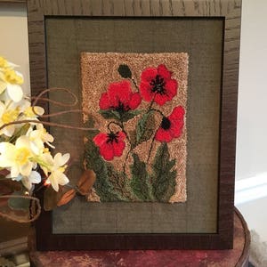 May include: A framed needlepoint artwork featuring a cluster of red poppies with green leaves and stems on a brown background.