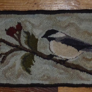 May include: A needlepoint rug depicting a small bird perched on a branch with red berries. The bird is brown, white, and black with a long tail. The background is a light beige with a swirling pattern.