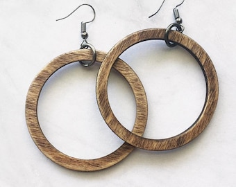 Puede incluir: Un par de pendientes circulares con un acabado de veta de madera natural en tono marrón claro. Cada pendiente está suspendido de un gancho de metal oscuro y una pequeña argolla, creando un diseño de aro sencillo con una textura visible.
