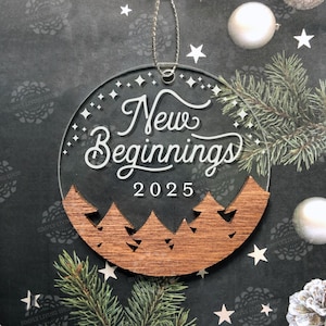 Puede incluir: Adorno navideño de acrílico transparente con las palabras "New Beginnings 2025" en escritura blanca. La parte inferior del adorno presenta un recorte de madera de pinos. El adorno cuelga de una cadena plateada y está rodeado de decoraciones navideñas.