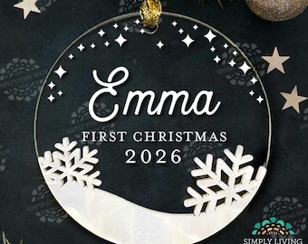 Puede incluir: Adorno navideño redondo de acrílico transparente con el nombre "Emma", el texto "FIRST CHRISTMAS 2026" y diseños de copos de nieve. El adorno tiene una cinta dorada para colgar. Estrellas y vegetación en el fondo.
