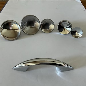 May include: A collection of polished chrome cabinet hardware. The set includes five circular knobs of varying sizes and a curved handle. The reflective surfaces create a bright, modern aesthetic, suitable for kitchen or bathroom cabinetry.