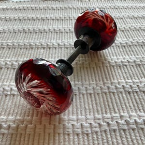 May include: A pair of round, deep red glass door knobs connected by a dark metal spindle. Each knob features a clear, starburst-like cut glass pattern radiating from the center, with additional circular cutouts around the outer edges of the glass.