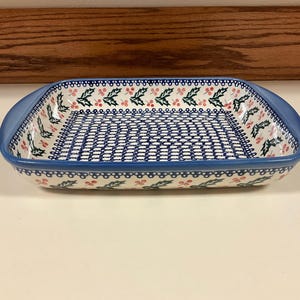 May include: A rectangular ceramic baking dish with rounded handles, featuring a blue rim and a decorative pattern of green holly leaves and red berries on a white background, with a blue and white geometric lattice design in the center.