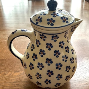 May include: A cream-colored ceramic pitcher with a matching lid, featuring a repeating pattern of small blue flowers with brown centers. The pitcher has a curved handle, a pointed spout, and a dark blue knob on the lid.