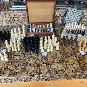 May include: A collection of multiple chess sets featuring black, white, and brown plastic or wooden chess pieces. The set includes a brown wooden storage box with internal compartments, a circular board, and a rectangular box labeled Chess.