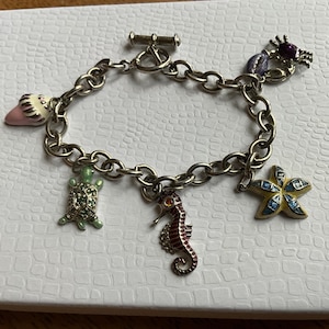 May include: A silver-toned metal charm bracelet featuring a toggle clasp and five distinct sea-themed charms: a pink and white seashell, a green sea turtle, a brown seahorse, a yellow and blue starfish, and a purple crab with a dark center stone.