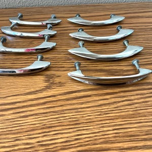 May include: Eight shiny, silver-toned cabinet pulls with curved handles and screw posts are arranged on a wooden surface. The pulls are evenly spaced, reflecting light and adding a touch of elegance.
