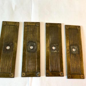 May include: Four antique brass rectangular plates with a central circular hole and vertical grooved texture. Each plate has four small square holes in the corners. The plates have a tarnished, aged appearance.