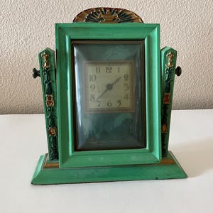 May include: A vintage green mantel clock with a rectangular face and a gold-colored sunburst design at the top. The clock face has black hands and gold numerals. The clock is made of metal and has decorative side panels.