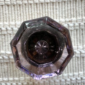 May include: A translucent, octagonal glass door knob with a light purple tint. The knob features a faceted, star-like pattern radiating from the center, with a smooth, polished surface and visible internal air bubbles within the glass structure.