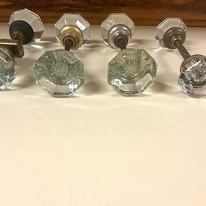 May include: A set of four clear glass door knobs with octagonal facets and starburst centers. Each knob features a metal base in varying finishes, including brass, silver, and bronze tones, attached to a metal spindle for door installation.