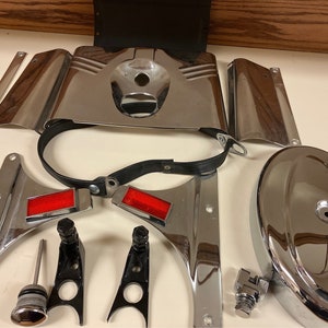 May include: A collection of chrome motorcycle parts, including fenders, brackets, and a cover, arranged on a light surface. The parts have a shiny, reflective finish, with red reflectors and black accents. The Harley-Davidson logo is visible on one of the parts.