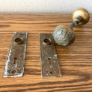 May include: A set of antique brass door hardware including two rectangular backplates with intricate embossed patterns and keyholes, paired with a matching round door knob featuring a detailed decorative design on its surface.