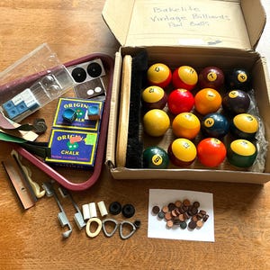 May include: A collection of vintage billiards accessories including a cardboard box labeled Bakelite Vintage Billiards Pool Balls containing sixteen numbered balls and a brush, a red plastic triangle, chalk boxes, cue tips, and small hardware.