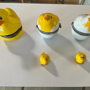May include: A collection of yellow and white ceramic trinket boxes and figurines. The set includes a duck-shaped box, two egg-shaped boxes with yellow chick details, and two small chick figurines. The items are arranged on a white surface.