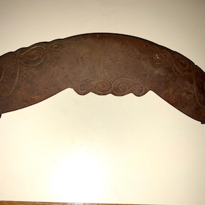 May include: A curved, rusty-brown metal object with decorative swirl patterns along the top edge. The object has a unique shape, possibly a decorative element or a tool, with a small projection at one end.