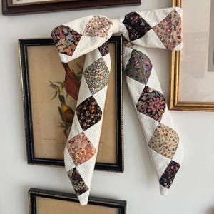 以下が含まれることがあります： キルト生地で作られた、大きな装飾用のリボン。リボンは、ピンク、茶色、緑など、さまざまな色の花柄生地のひし形のパッチが付いた白いベースが特徴です。白い壁に掛けられており、背景には額入りのアートワークがあります。