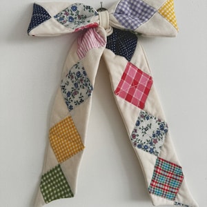 以下が含まれることがあります： クリーム色の生地で作られた、大きな装飾用のリボン。カラフルなひし形のパッチで飾られています。パッチには、花柄、ギンガムチェック、水玉模様など、さまざまなパターンがあります。白い壁に掛けられています。