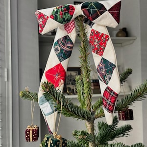 以下が含まれることがあります： 赤、緑、白の生地を使ったパッチワークデザインのキルトクリスマスツリーリボン。リボンはひし形の模様で飾られ、小さな常緑樹に飾られています。小さなギフトボックスのオーナメントが木から吊り下げられています。