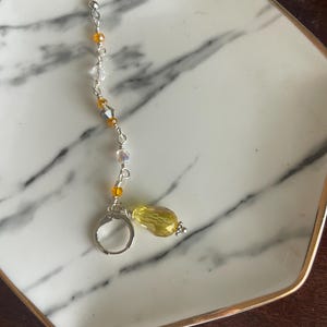 May include: A single earring with a silver hook and a dangling design. The earring features a series of small, clear and amber-colored beads strung along a silver wire. A large, faceted yellow bead hangs at the bottom, adding a pop of color.