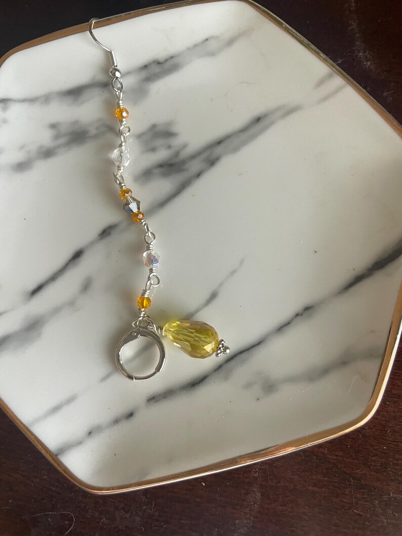 May include: A single earring with a silver hook and a dangling design. The earring features a series of small, clear and amber-colored beads strung along a silver wire. A large, faceted yellow bead hangs at the bottom, adding a pop of color.