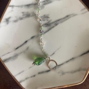May include: A single, dangling earring with a silver hook and a series of clear and green beads. The earring features a large, faceted green teardrop-shaped bead at the bottom. The earring is displayed on a white and gray marble-patterned plate.
