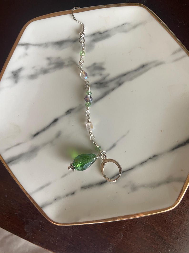 May include: A single, dangling earring with a silver hook and a series of clear and green beads. The earring features a large, faceted green teardrop-shaped bead at the bottom. The earring is displayed on a white and gray marble-patterned plate.