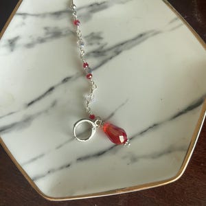 May include: A single earring with a silver hook and a dangling red teardrop-shaped bead. The earring features a delicate chain with small red and clear beads. The earring is resting on a white and gray marble patterned tray with a gold rim.