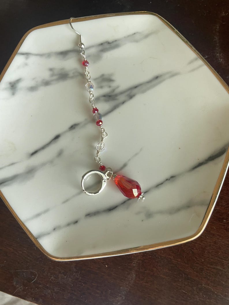 May include: A single earring with a silver hook and a dangling red teardrop-shaped bead. The earring features a delicate chain with small red and clear beads. The earring is resting on a white and gray marble patterned tray with a gold rim.