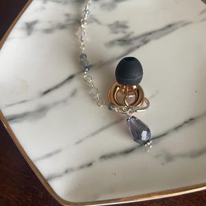 May include: Earring with a silver hook and a chain of small, clear and blue beads. The earring features a gold-colored ring and a black earbud-shaped element, with a teardrop-shaped purple crystal pendant.