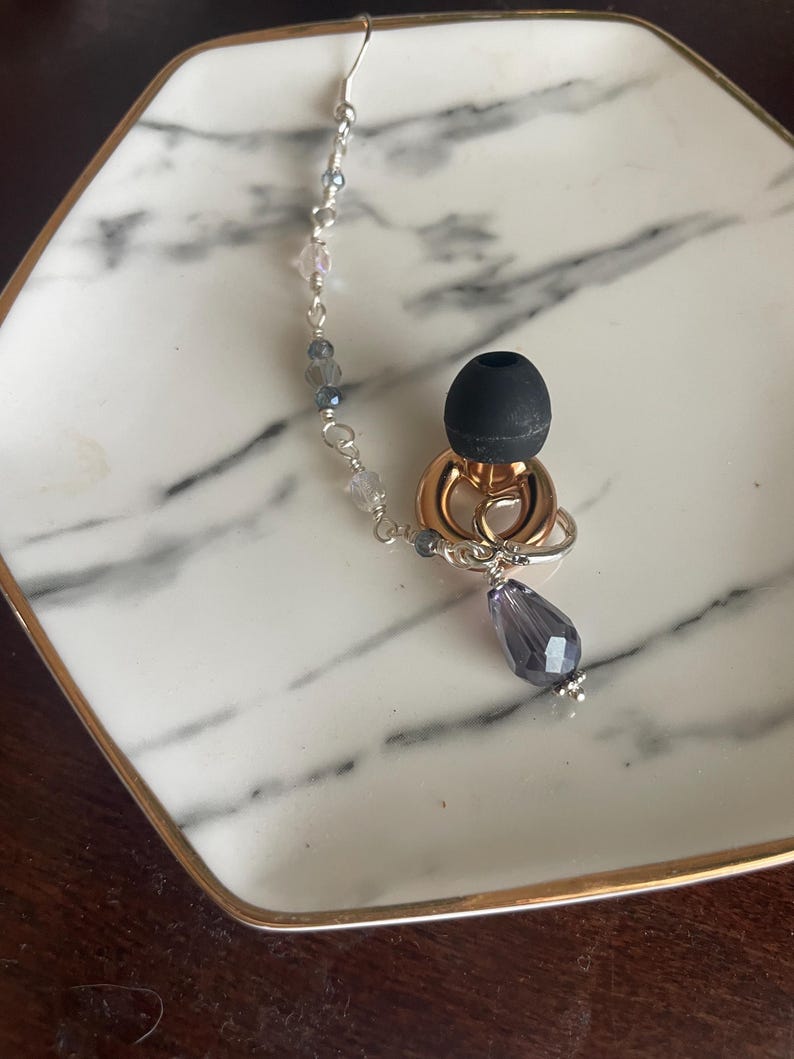 May include: Earring with a silver hook and a chain of small, clear and blue beads. The earring features a gold-colored ring and a black earbud-shaped element, with a teardrop-shaped purple crystal pendant.
