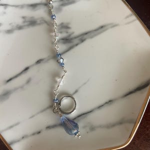May include: A silver earring with a dangling design. The earring features a series of small, clear and blue beads strung along a silver chain, culminating in a large, teardrop-shaped blue crystal. A small silver ring is attached.