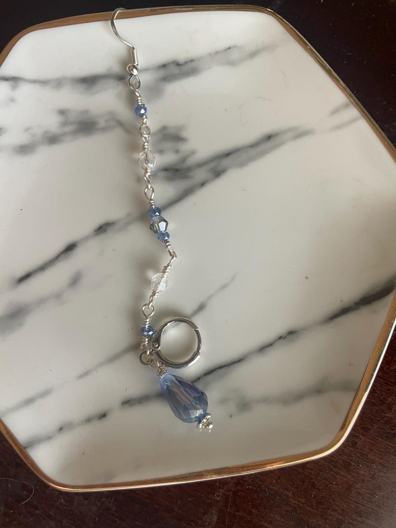 May include: A silver earring with a dangling design. The earring features a series of small, clear and blue beads strung along a silver chain, culminating in a large, teardrop-shaped blue crystal. A small silver ring is attached.