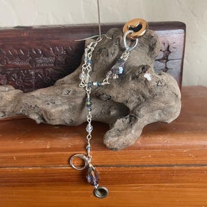 May include: A handmade earring with a silver hook and a dangling chain of small, clear and blue beads. The earring features a large, teardrop-shaped purple bead and a gold-colored ring. A needle is inserted through the earring.