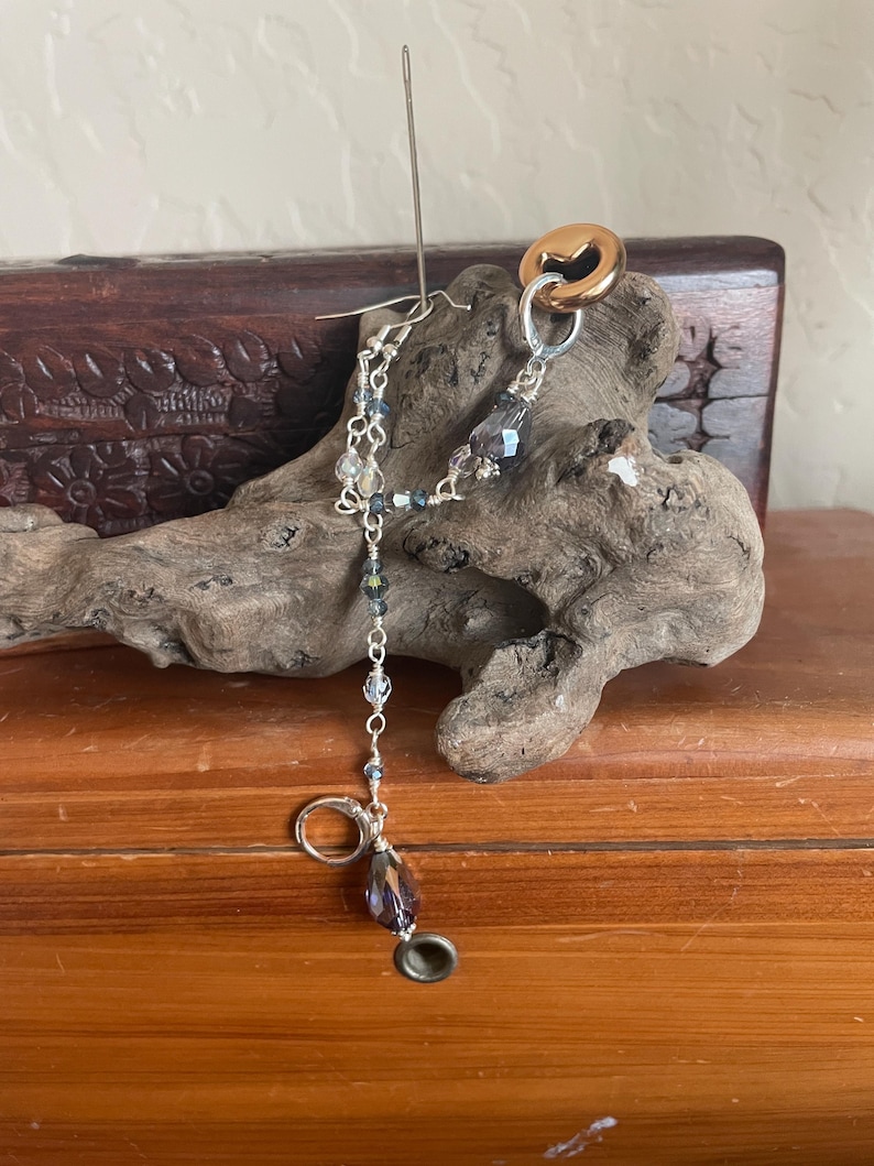 May include: A handmade earring with a silver hook and a dangling chain of small, clear and blue beads. The earring features a large, teardrop-shaped purple bead and a gold-colored ring. A needle is inserted through the earring.