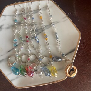 May include: A collection of colorful beaded earrings displayed on a white and gold-rimmed plate. Each earring features a silver hook, a chain of small beads, and a larger, teardrop-shaped crystal in various colors, including blue, green, red, and yellow.