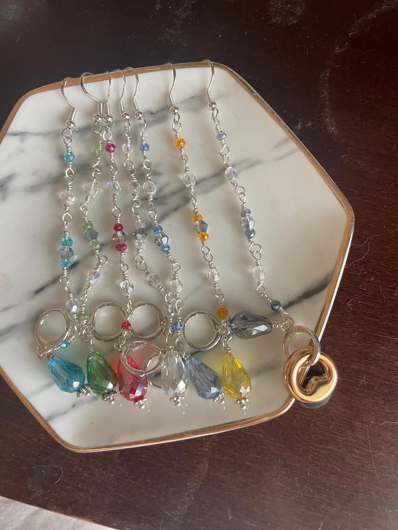 May include: A collection of colorful beaded earrings displayed on a white and gold-rimmed plate. Each earring features a silver hook, a chain of small beads, and a larger, teardrop-shaped crystal in various colors, including blue, green, red, and yellow.