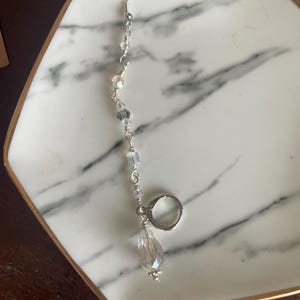 May include: A single silver earring with a clear, teardrop-shaped crystal pendant. The earring features a series of small, iridescent beads strung along a silver wire, culminating in a larger, faceted crystal. The earring is displayed on a white and gray marble tray.