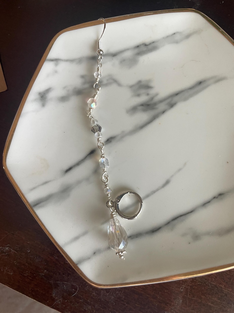May include: A single silver earring with a clear, teardrop-shaped crystal pendant. The earring features a series of small, iridescent beads strung along a silver wire, culminating in a larger, faceted crystal. The earring is displayed on a white and gray marble tray.
