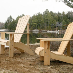 Pu&ograve; includere: Due sedie Adirondack in legno su una spiaggia sabbiosa vicino a un lago. Le sedie sono in legno naturale e hanno un design classico.