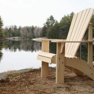 Pu&ograve; includere: Una sedia Adirondack in legno con finitura naturale si trova su una spiaggia sabbiosa vicino a un lago. La sedia &egrave; realizzata in legno grezzo e ha uno schienale alto e braccioli.