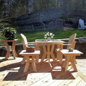 May include: A wooden outdoor dining set with a square table and four chairs. The table and chairs are made of natural wood and have a rustic look. The set is placed on a wooden deck with a large rock formation in the background.