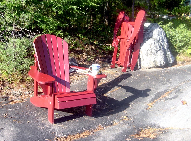 Puede incluir: Dos sillas Adirondack de madera roja, una con una mesa peque&ntilde;a y una taza encima, est&aacute;n sentadas en un camino de grava en un &aacute;rea boscosa.
