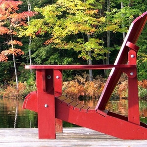 Puede incluir: Una silla Adirondack de madera roja se encuentra en un muelle de madera con vistas a un lago. La silla est&aacute; hecha de madera resistente y tiene un dise&ntilde;o c&oacute;modo. El lago est&aacute; rodeado de &aacute;rboles con follaje oto&ntilde;al.