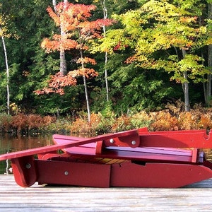 Puede incluir: Una silla plegable de madera roja est&aacute; sentada en un muelle de madera. La silla est&aacute; plegada y el respaldo est&aacute; apoyado contra un &aacute;rbol. El muelle est&aacute; sobre un lago con &aacute;rboles en el fondo.